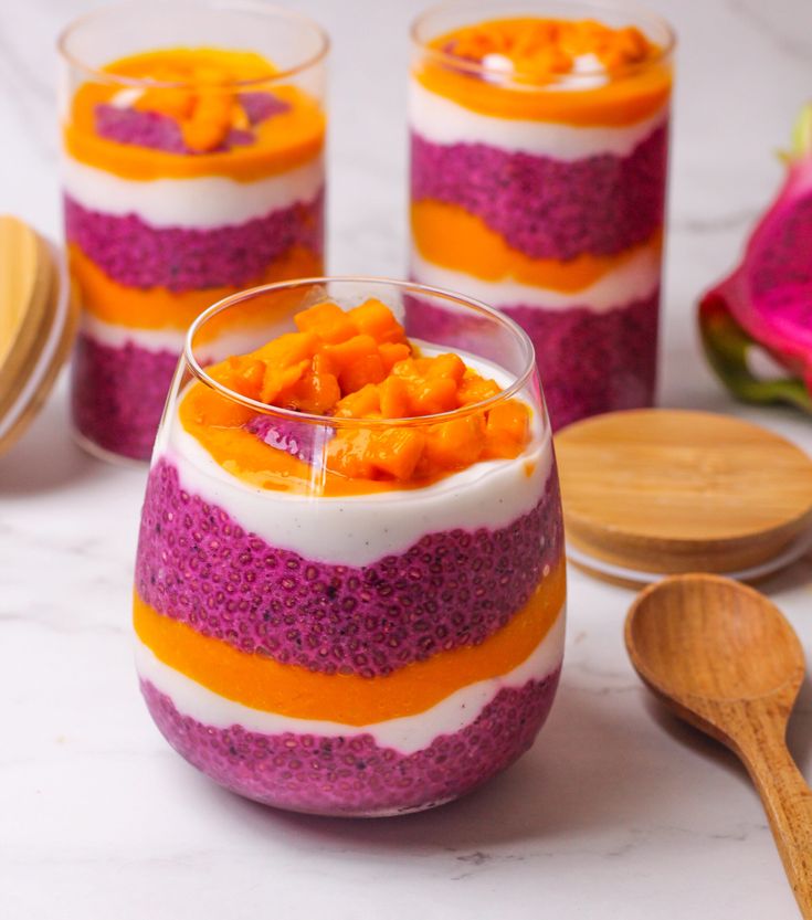 three glasses filled with colorful desserts on top of a white counter next to wooden spoons