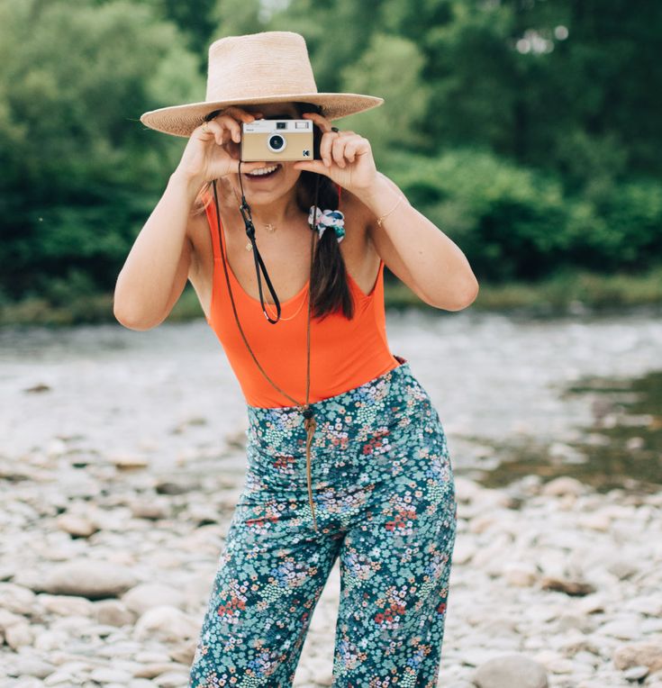 a woman in an orange tank top and floral pants is holding a camera up to her face