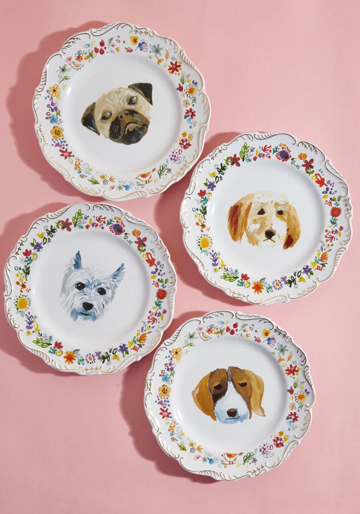 four plates with dogs painted on them sitting on a pink tableclothed surface in front of a pink wall