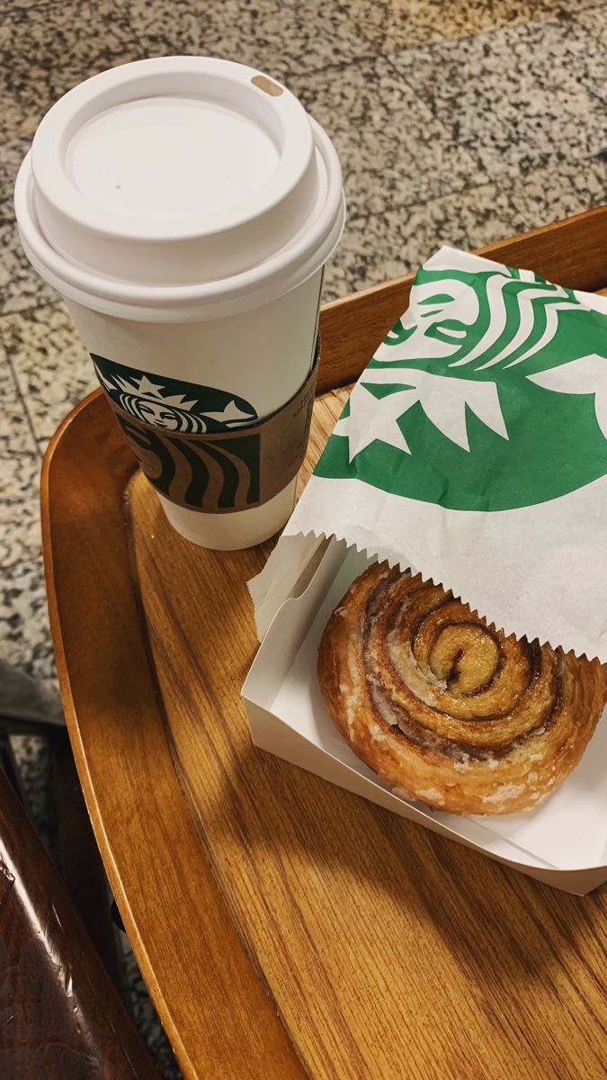 a cup of coffee sitting on top of a wooden tray next to a bag with cinnamon rolls