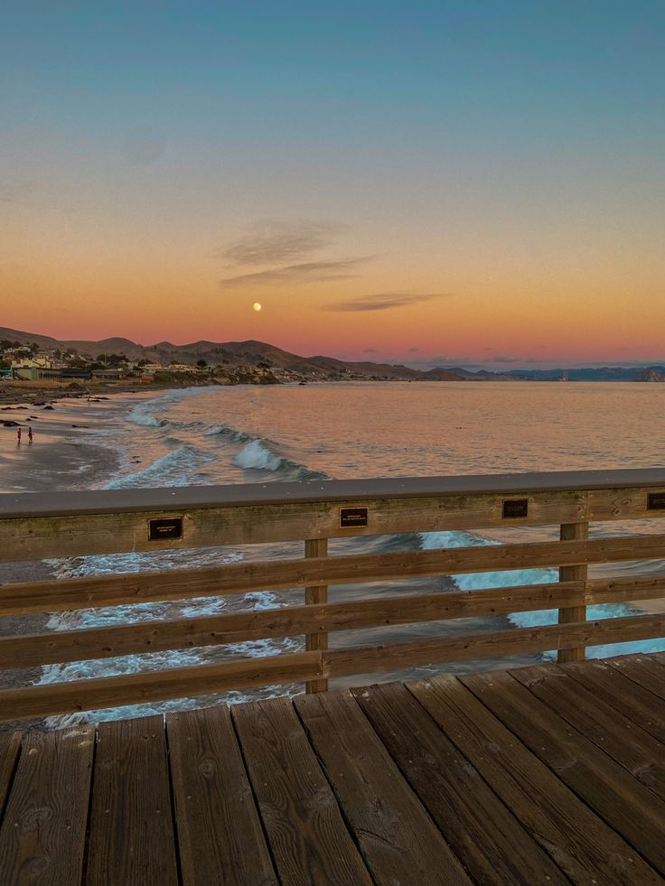 the sun is setting over the ocean and beach as people walk on the boardwalk near the water