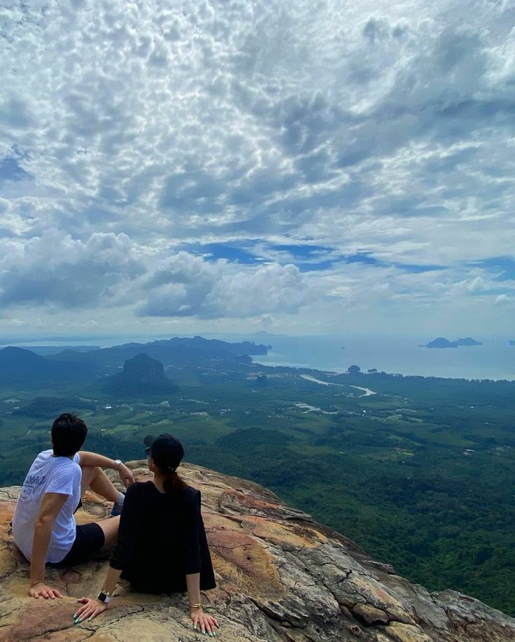 two people sitting on the edge of a cliff looking out at mountains and clouds in the distance