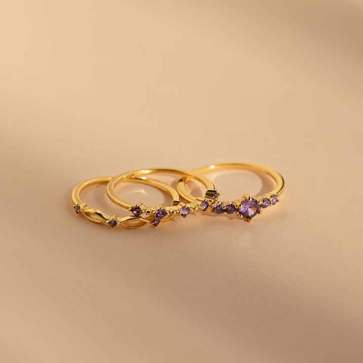 Amethyst is believed to bring about serenity and peace to those who wear it. Our Amethyst Stacking Ring Set not only can inspire those qualities, but also will make a fashion forward statement. The delicate purple gemstones in each of the three rings sparkle, letting you be in the spotlight where you are meant to be. Material: High Quality Solid 925 Sterling Silver Finish: 18K Gold Featuring a Set of 3 Amethyst CZ Gemstone Rings ($75 value): ~1mm Sprinkle Band with ~2mm Stones ~1mm Open Oval Ban Gold Dainty Amethyst Ring, Gold Amethyst Ring In Dainty Style, Gold Amethyst Birthstone Promise Ring, Gold Amethyst Ring For Promise, Gold Amethyst Birthstone Ring For Promise, Gold Amethyst Stackable Rings, Gold Amethyst Stackable Ring Fine Jewelry, Gold Amethyst Crystal Ring With Gemstone, Gold Amethyst Ring With Spiritual Style