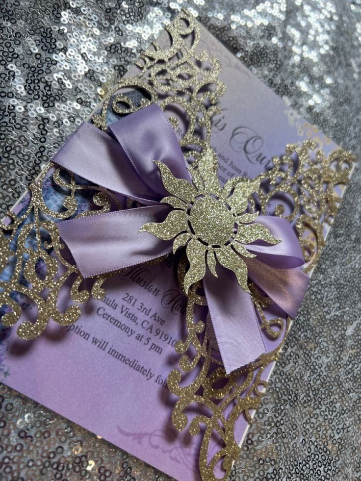 a purple and gold wedding card with a large bow on the front, sitting on top of glittery fabric