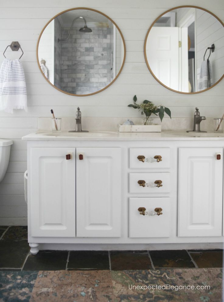 two round mirrors are above the double sink vanity in this white bathroom with brick flooring