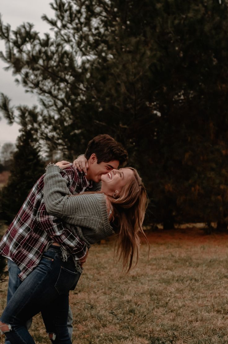 a man and woman are hugging in the grass with trees in the back ground behind them