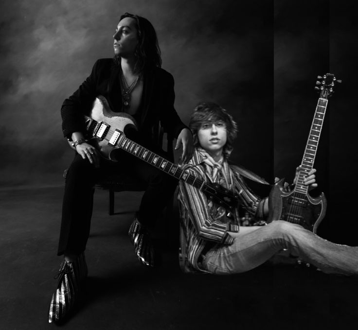 two young men sitting next to each other with guitars in their hands and one holding an electric guitar