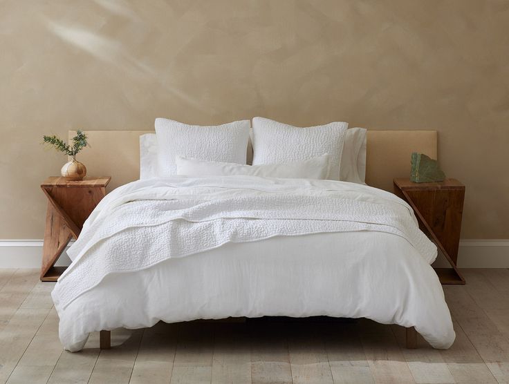 a bed with white linens and pillows in a neutral colored room, next to a wooden night stand