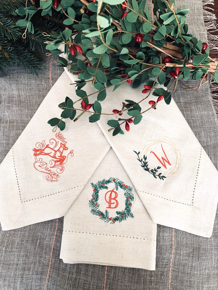 three napkins with embroidered monogrammed designs on them sitting next to some greenery