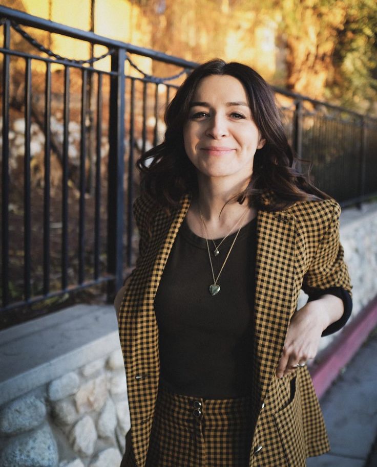 a woman standing next to a fence wearing a black top and yellow plaid blazer