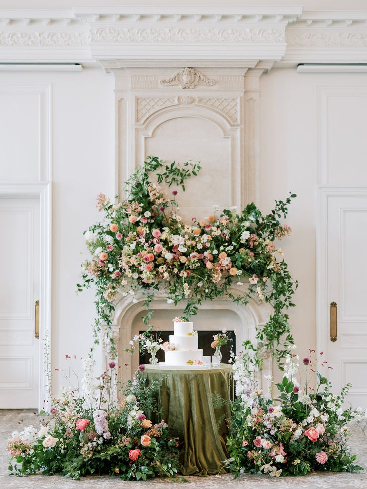 a wedding cake sitting on top of a table surrounded by flowers and greenery in front of a fireplace