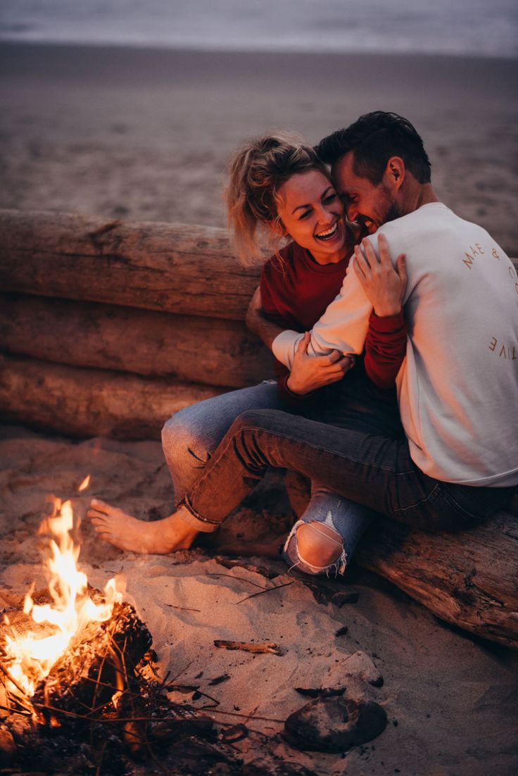 a man and woman sitting next to a fire on the beach