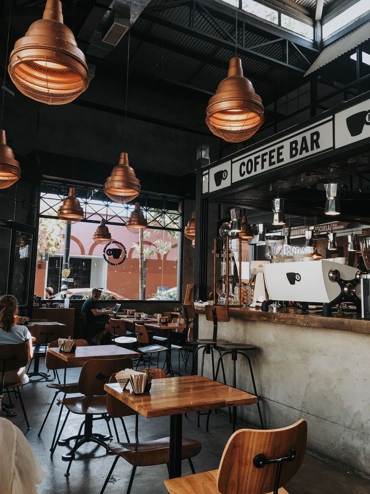 the interior of a coffee shop with wooden tables and chairs, hanging lights, and large windows