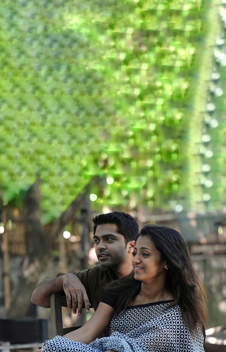 a man and woman sitting next to each other in front of a green plant covered wall