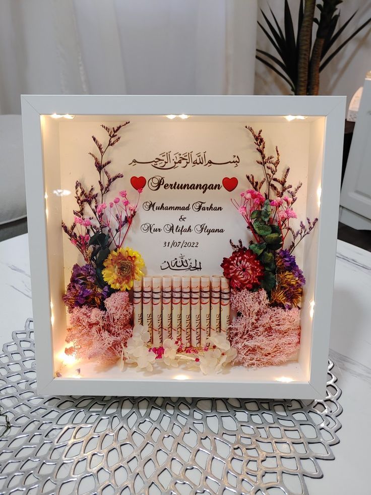 a white frame with flowers and lights in it on a table next to a potted plant