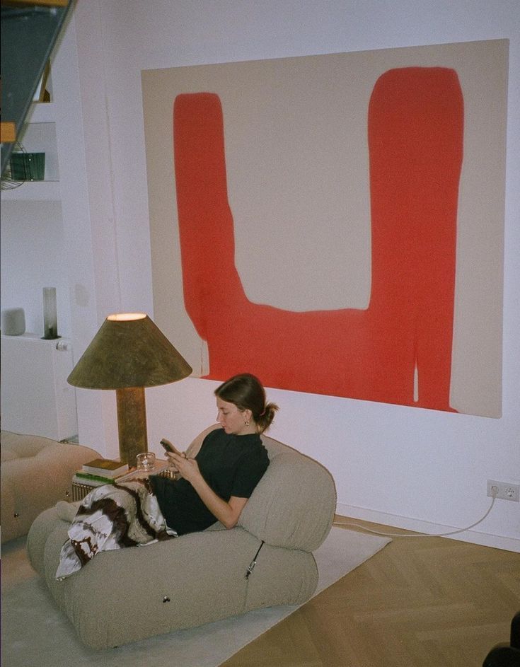 a woman sitting on a couch in front of a large red piece of art with a lamp next to her