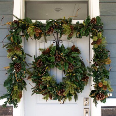 a wreath with pine cones and green leaves hanging on the front door to a house