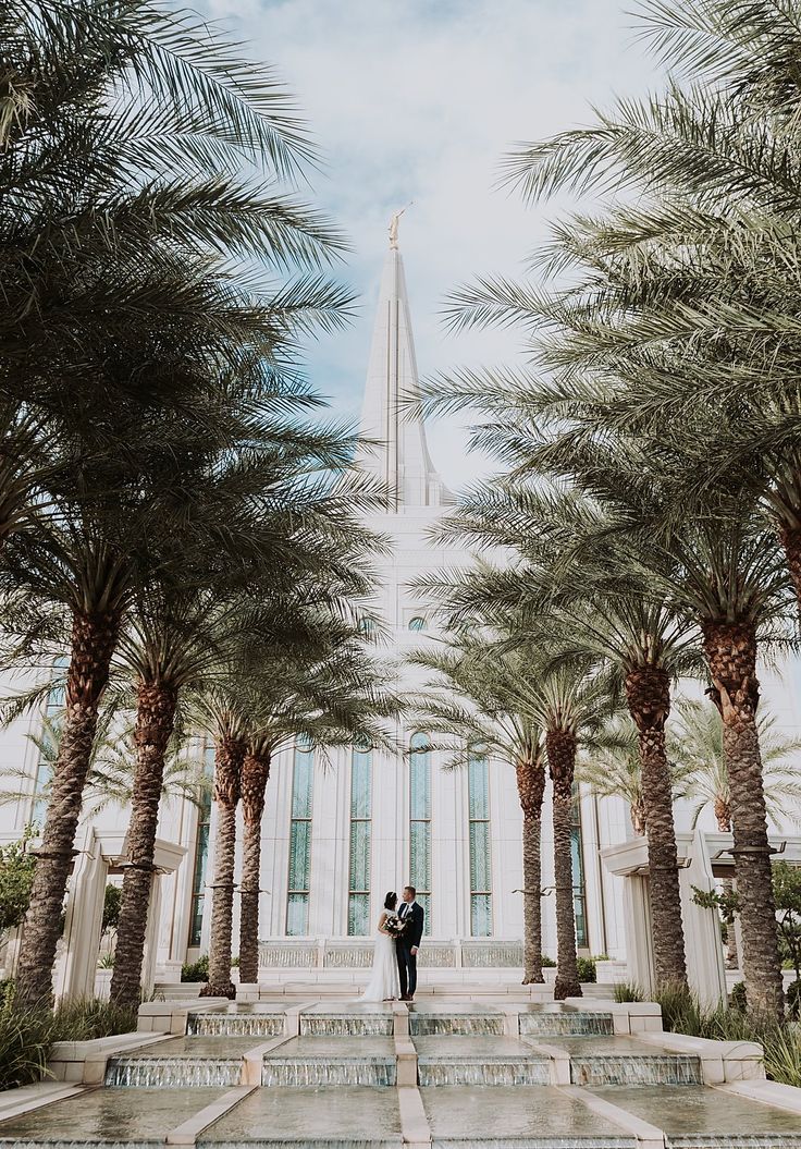 a bride and groom standing under palm trees in front of a tall building with a steeple