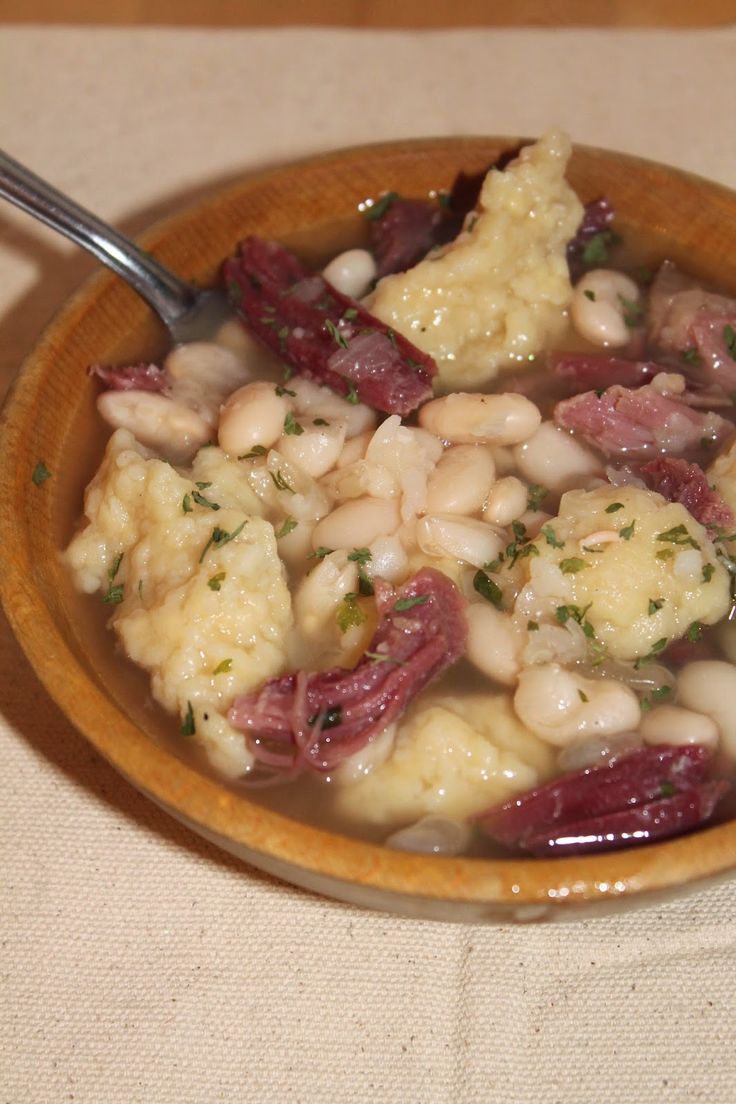 a bowl filled with meat and beans on top of a white table cloth next to a spoon