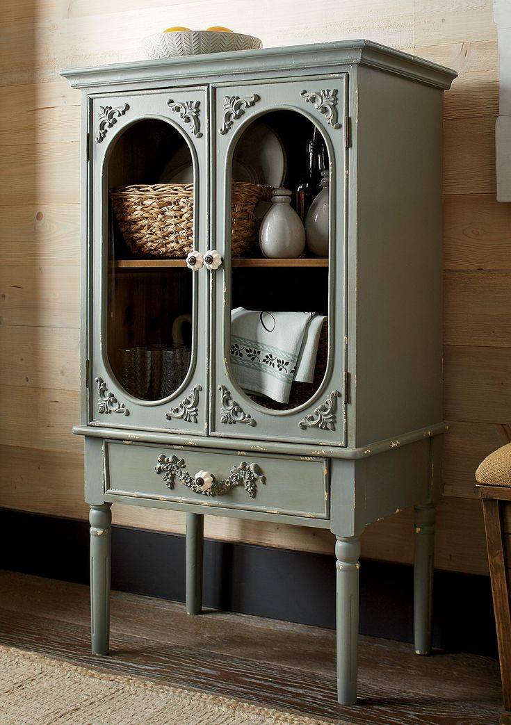 a green cabinet with two glass doors and some baskets on top of it, in front of a wooden wall