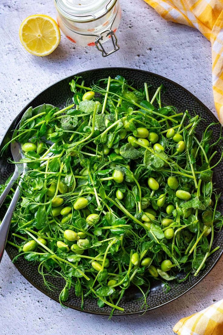 a black plate topped with green vegetables next to a jar of lemons and a spoon