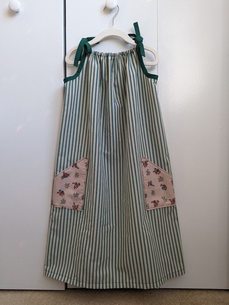 "Your child will feel cool and comfortable all summer long in this loose fitting sundress with adjustable tie straps. This dress features two large floral pockets on the front of the dress, and a hunter green striped pattern. This listing is for the green striped sundress as shown in the thumbnail. The blue dress on the model is only to show the fit of the dress. This dress is listed as size 5-7, but please be sure to check the measurements, and keep in mind that the neckline is elastic and the Green Summer Sundress For Picnic, Green Summer Sundress For Picnics, Green Cotton Sundress, Green Sundress With Tie Straps For Garden Party, Green Sundress With Adjustable Straps, Green Sundress For Picnic, Green Cotton Dresses With Adjustable Straps, Green Cotton Dress With Adjustable Straps, Green Summer Sundress With Adjustable Straps