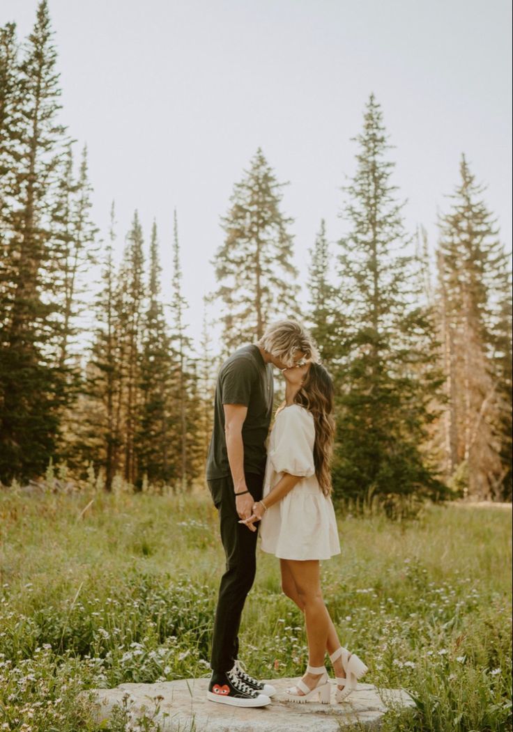 a man and woman standing on top of a rock in the grass with trees behind them