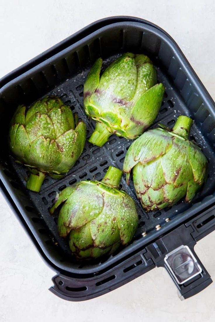 four green artichokes in a black container on a white counter top,