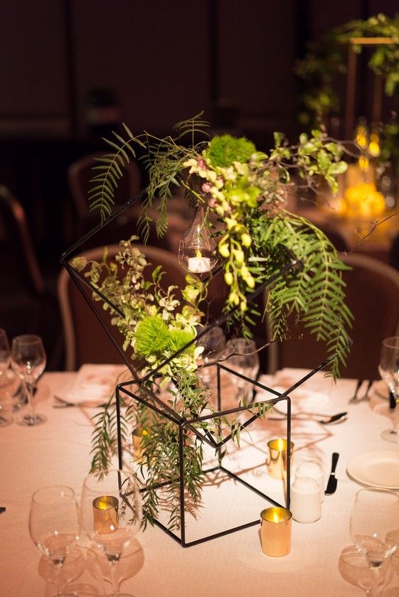 an arrangement of greenery is displayed in a geometric glass container on a table set with wine glasses and candles