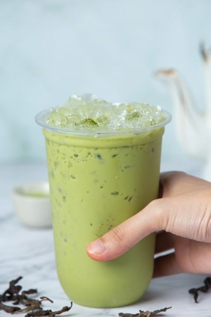 a hand is holding a green drink with ice and tea on the table next to it