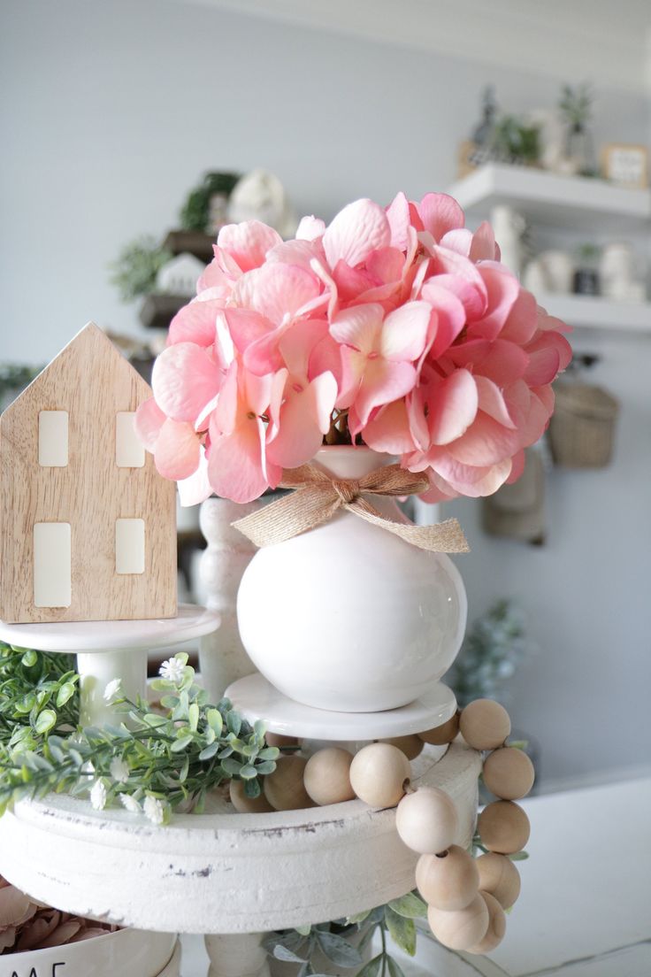 a white vase with pink flowers in it on top of a table next to a house