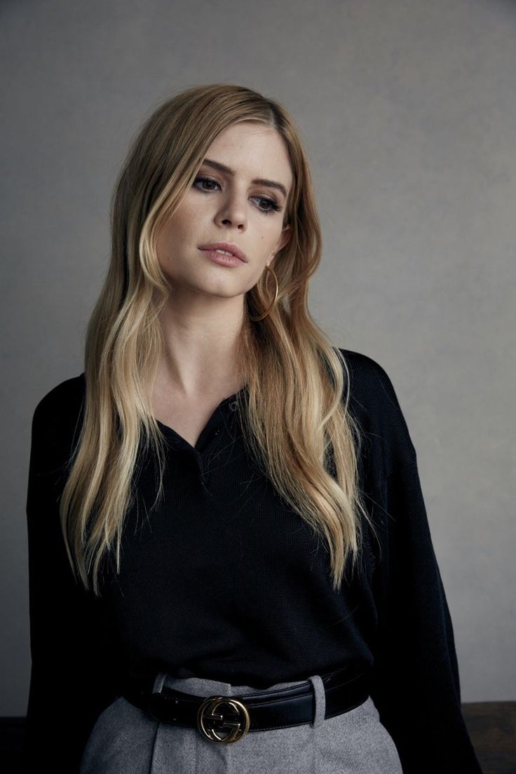 a woman with long blonde hair wearing a black shirt and grey pants, standing in front of a gray wall