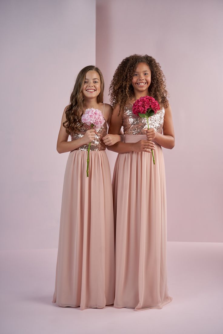 two girls in long dresses holding flowers and smiling at the camera with one girl standing next to her