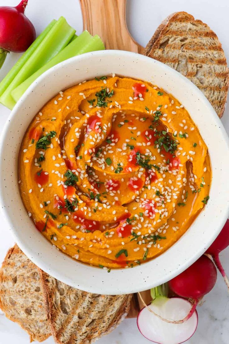 a white bowl filled with carrot hummus next to sliced bread and celery