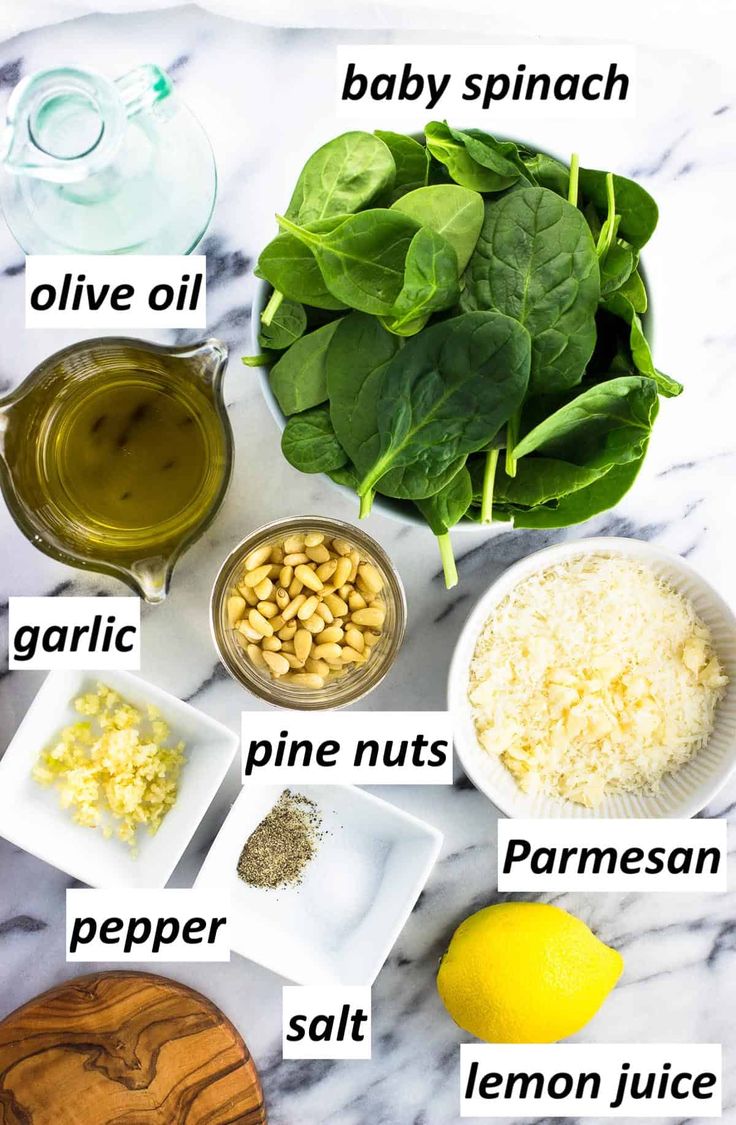 ingredients to make spinach salad laid out on a marble counter top with lemons, olive oil, pine nuts, parmesan cheese and baby spinach