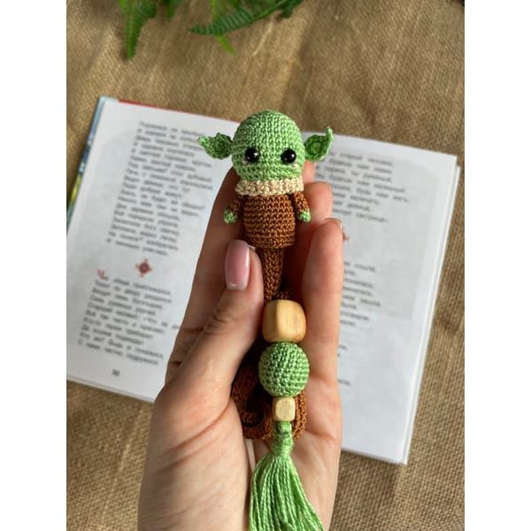 a hand holding a small crocheted yoda star wars doll next to an open book