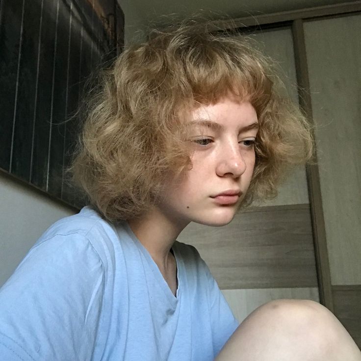 a young woman with curly hair sitting on the floor looking at her cell phone while wearing a blue shirt