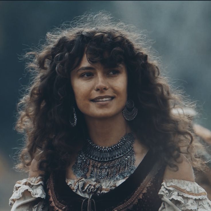 a woman with long curly hair wearing a brown dress and earrings, standing in front of a horse