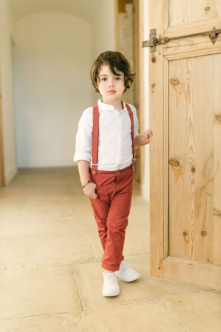 a little boy standing in front of a door wearing suspenders and a white shirt