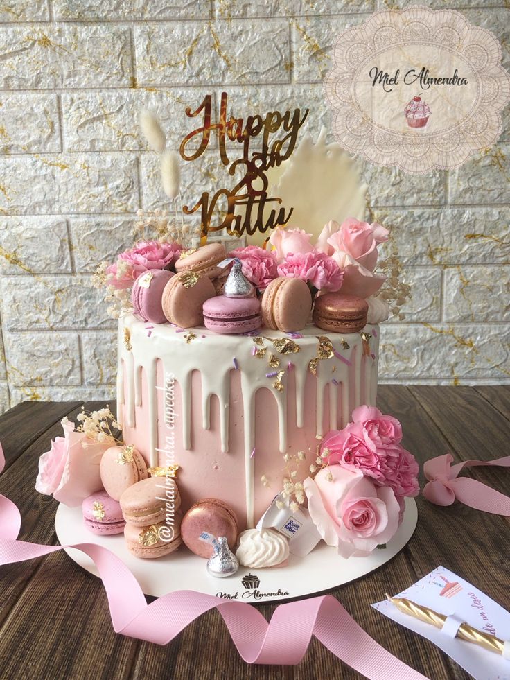 a birthday cake decorated with pink flowers and macaroons on a wooden table next to a brick wall