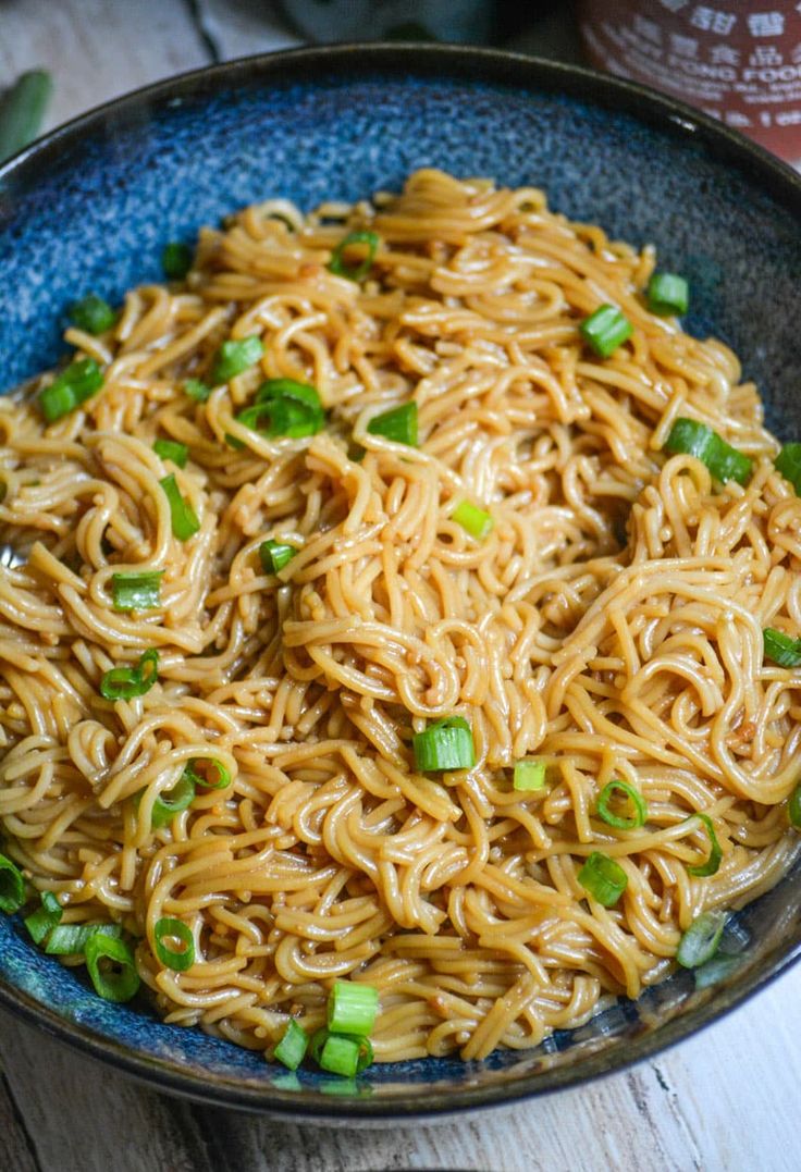 a blue bowl filled with noodles and green onions on top of a wooden table next to a jar of ketchup