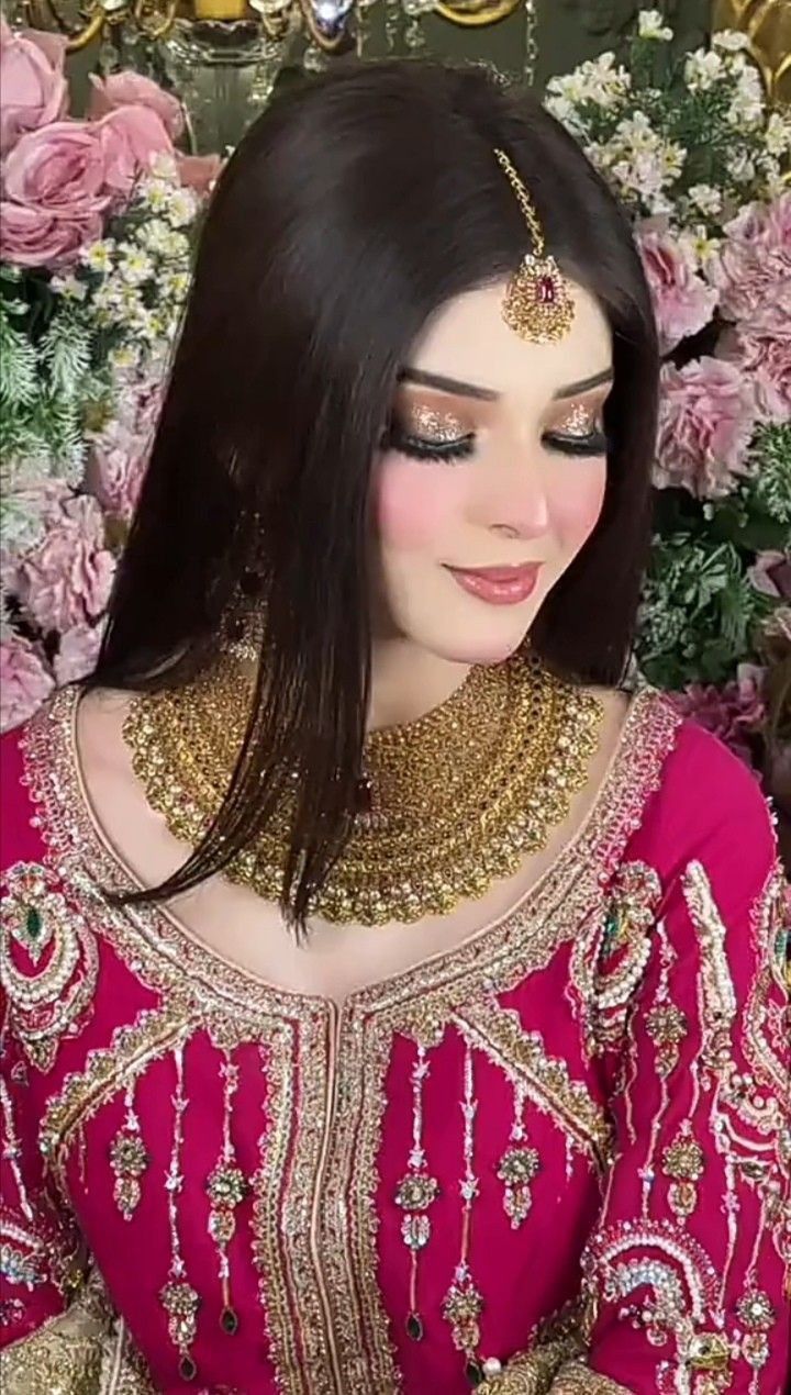 a woman wearing a pink outfit with gold jewelry and flowers in the background, looking down at her phone