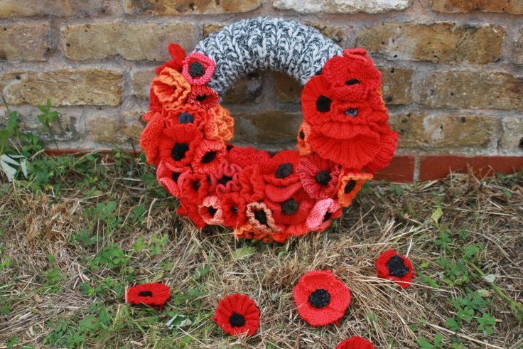 a knit wreath with red flowers on the grass next to a brick wall and some straw