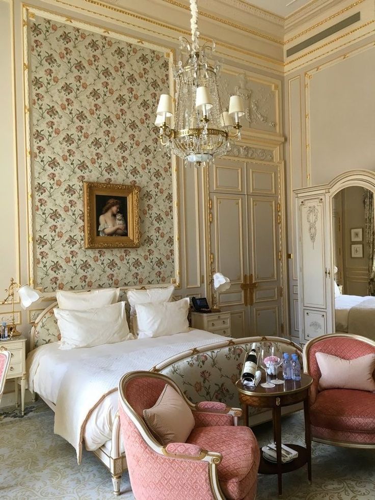 a bedroom with two pink chairs in front of a white bed and chandelier