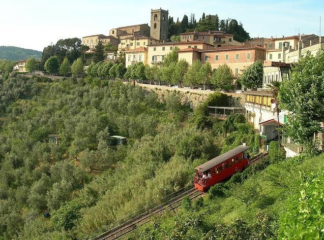 a red train traveling down tracks next to a lush green hillside covered in trees and buildings