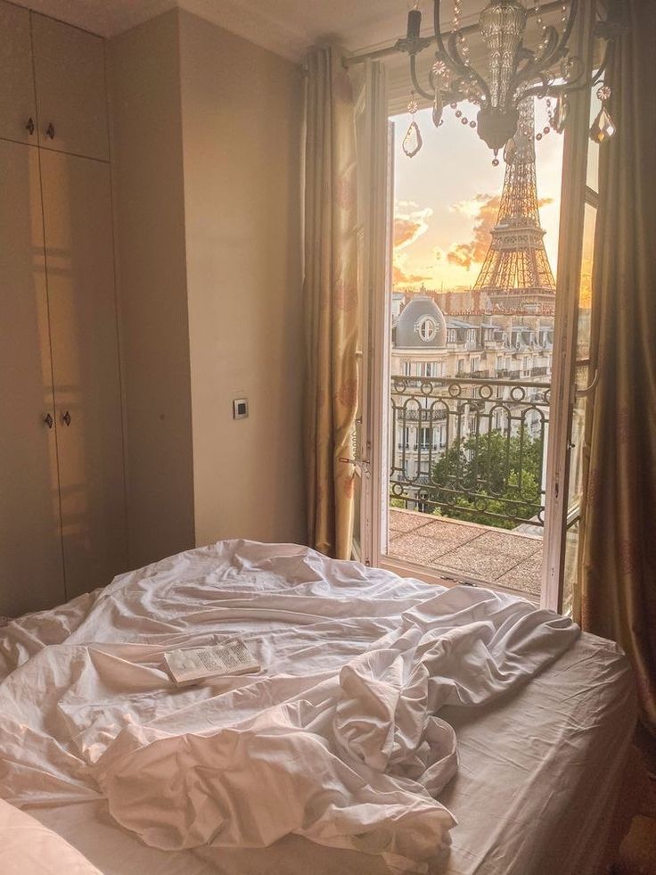 an unmade bed with the eiffel tower in the background from a bedroom window