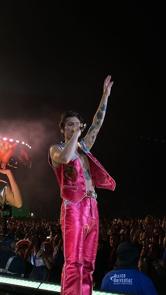 a man in pink outfit standing on stage with his hand up to the sky and fireworks behind him