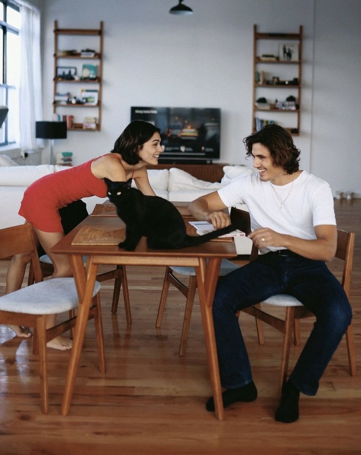 a man and woman sitting at a table with a black cat on it's lap