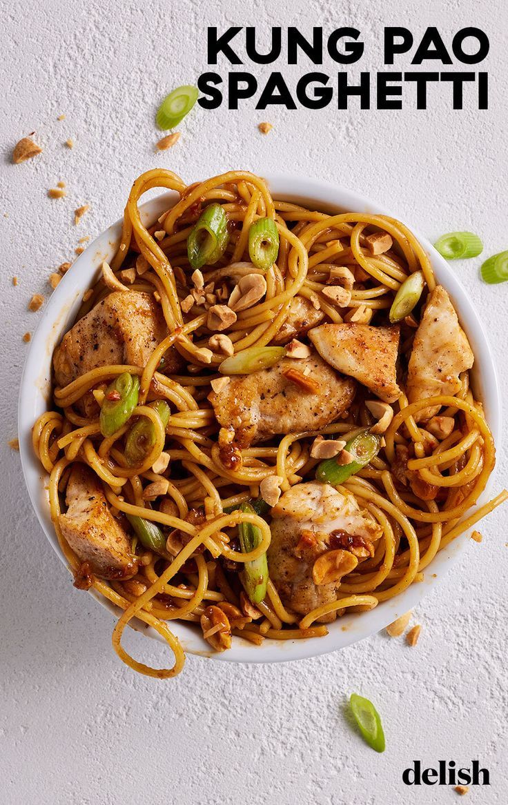 a white bowl filled with noodles and chicken on top of a table next to cashews