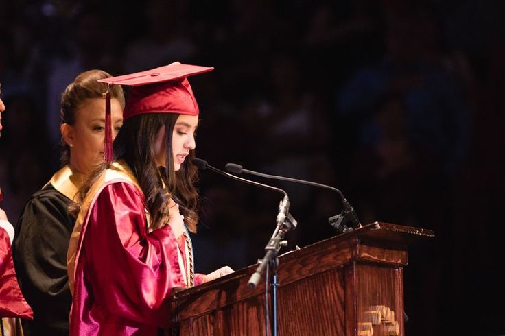 two girls in graduation gowns stand at a podium and speak into a microphone while another girl stands behind them